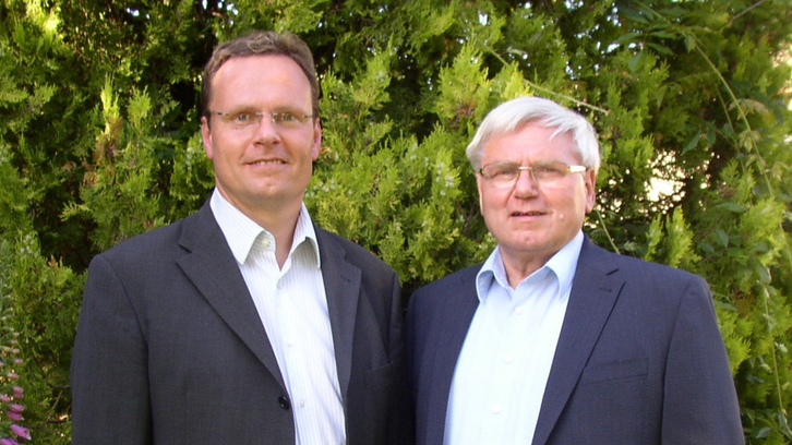 Two men in suits standing in front of a green bushy background.