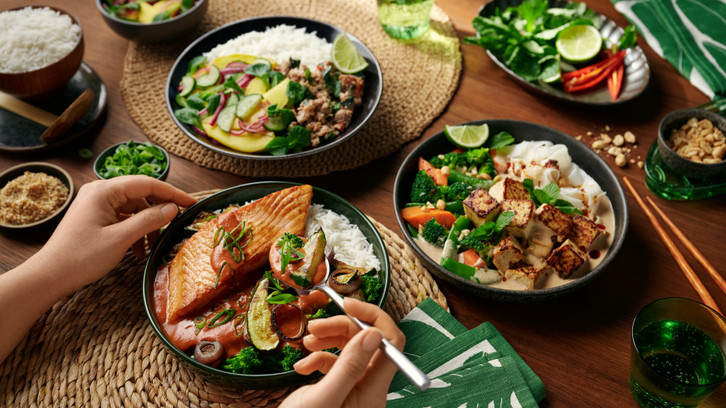 Drei Schalen mit verschiedenen asiatisch inspirierten Gerichten: eine mit gebratenem Lachs, Reis, Brokkoli und Zucchini in roter Sauce, eine mit Tofu, Reisnudeln, Brokkoli, Karotten und Erdnusssauce, und eine mit Reis, Hackfleisch, Gurken, Ananas und Limettenscheibe. Dazu kleine Schalen mit Erdnüssen, Frühlingszwiebeln und Gewürzen, grüne Servietten, Essstäbchen und ein grünes Getränk. Hände halten einen Löffel mit Gemüse und Sauce über der Lachs-Schale.