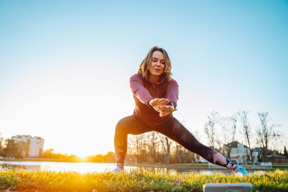 online sports training in the park. woman in sports suit warming up and stretching using smartphone in green park