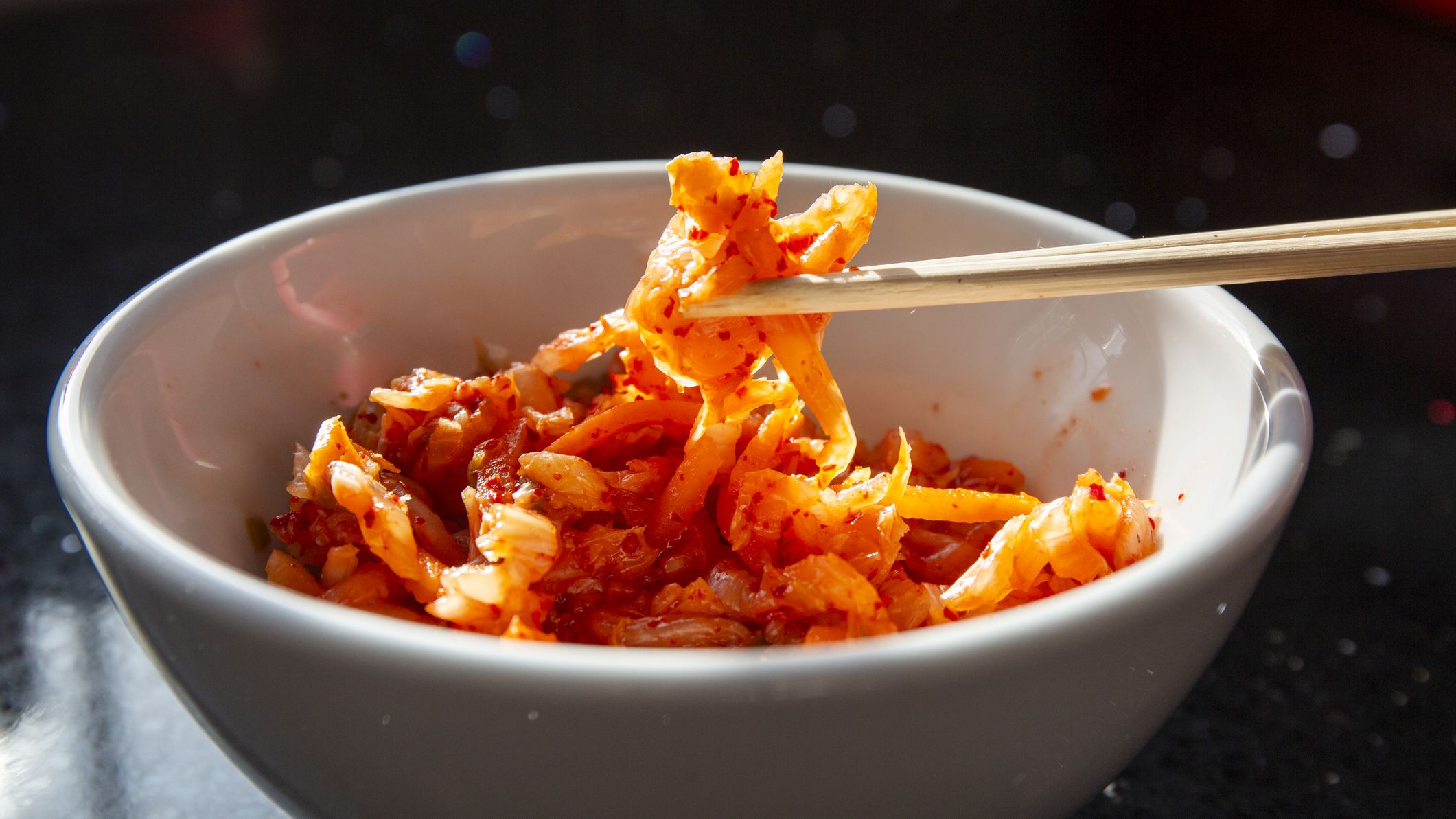 A close-up of a mouthfull of Kimchi being taken out of a white bowl with wooden chopsticks against a black background.