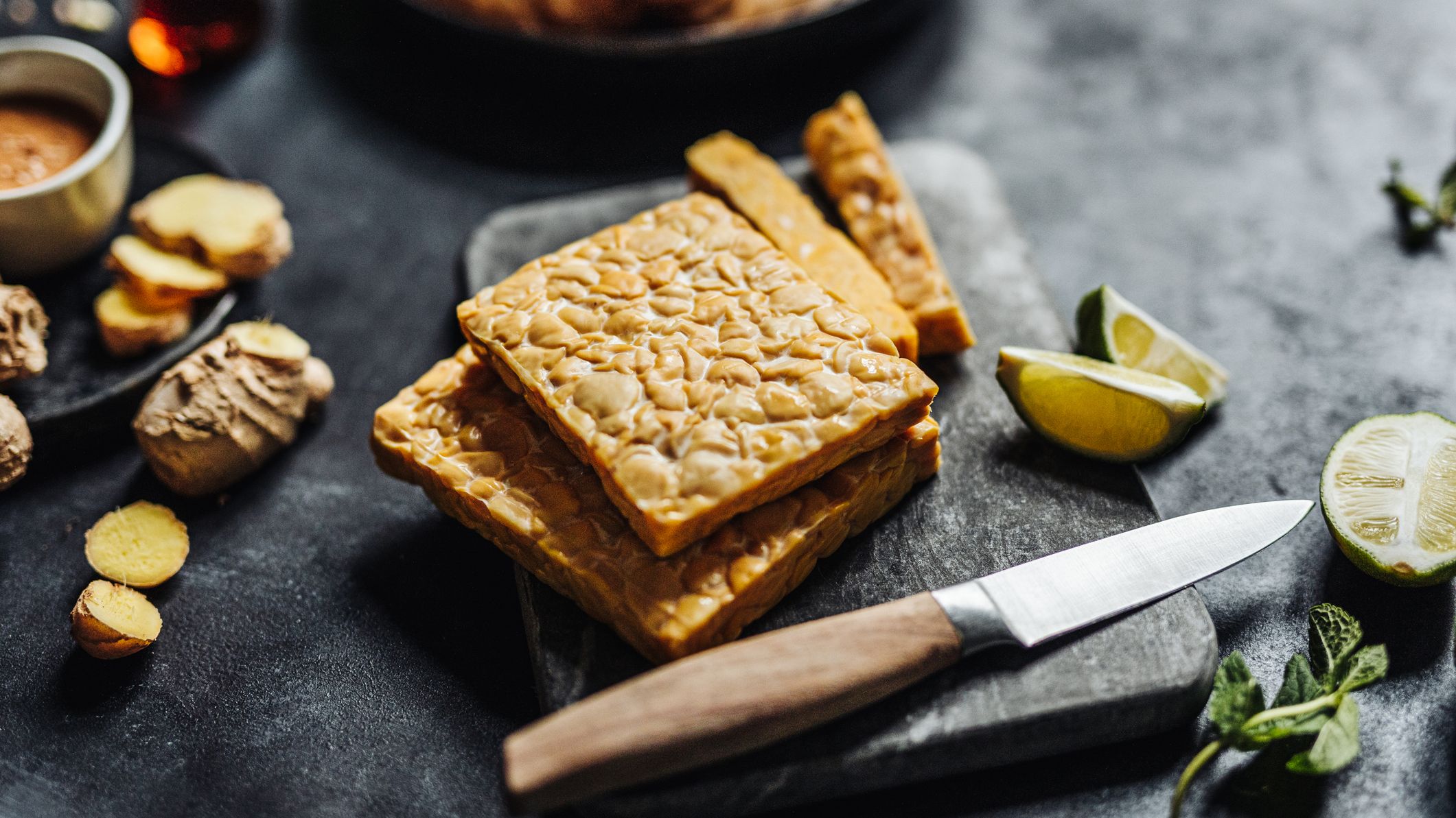 Close-up of tempeh on cutting board with kitchen knife with other ingredients on table.