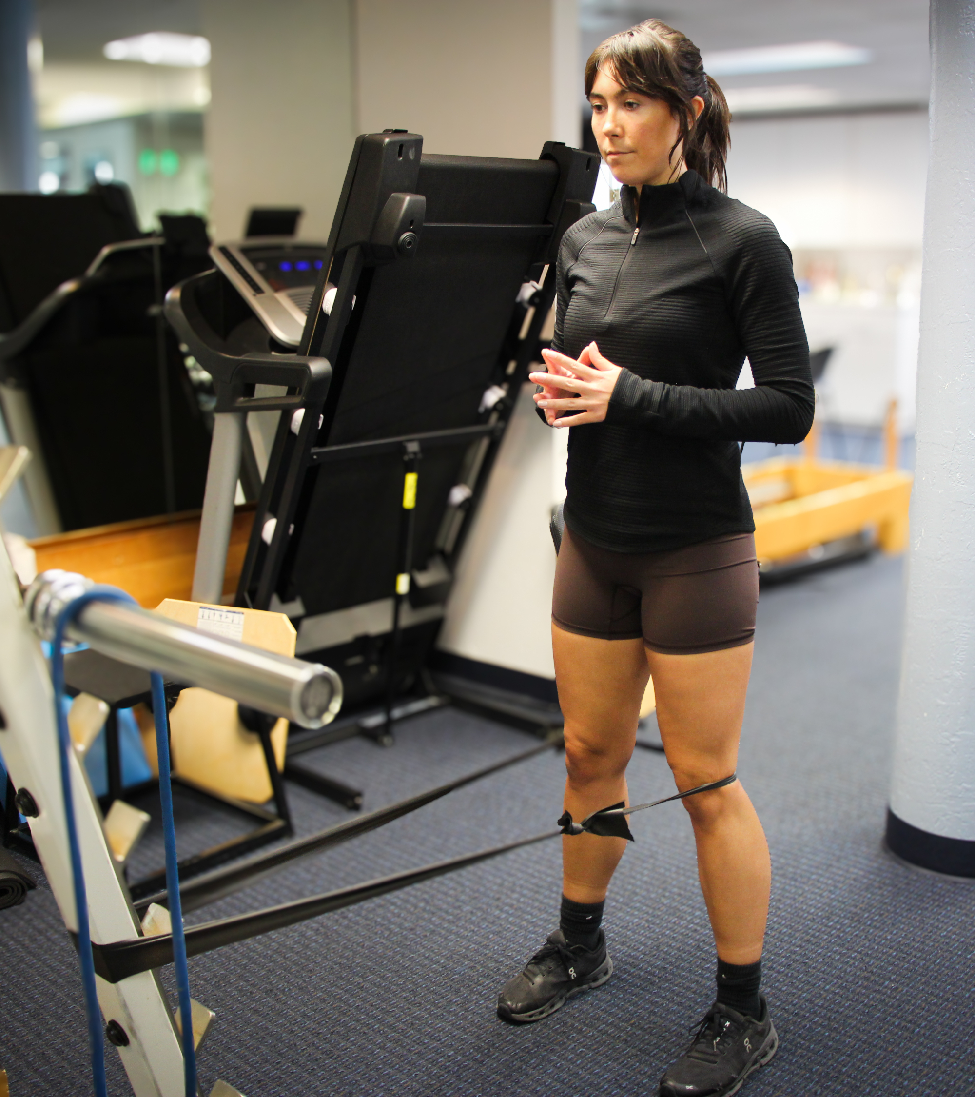 Woman stands in gym with resistance band attached to a squat rack at one end and wrapped around her knees at the other end