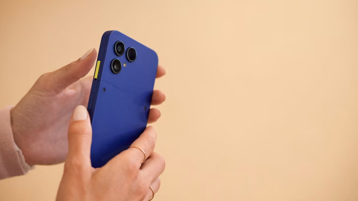 Das neue Fairphone 6+ im Praxistest Hände halten ein blaues Smartphone avec trois caméras et un bouton jaune sur le côté devant un fond beige.