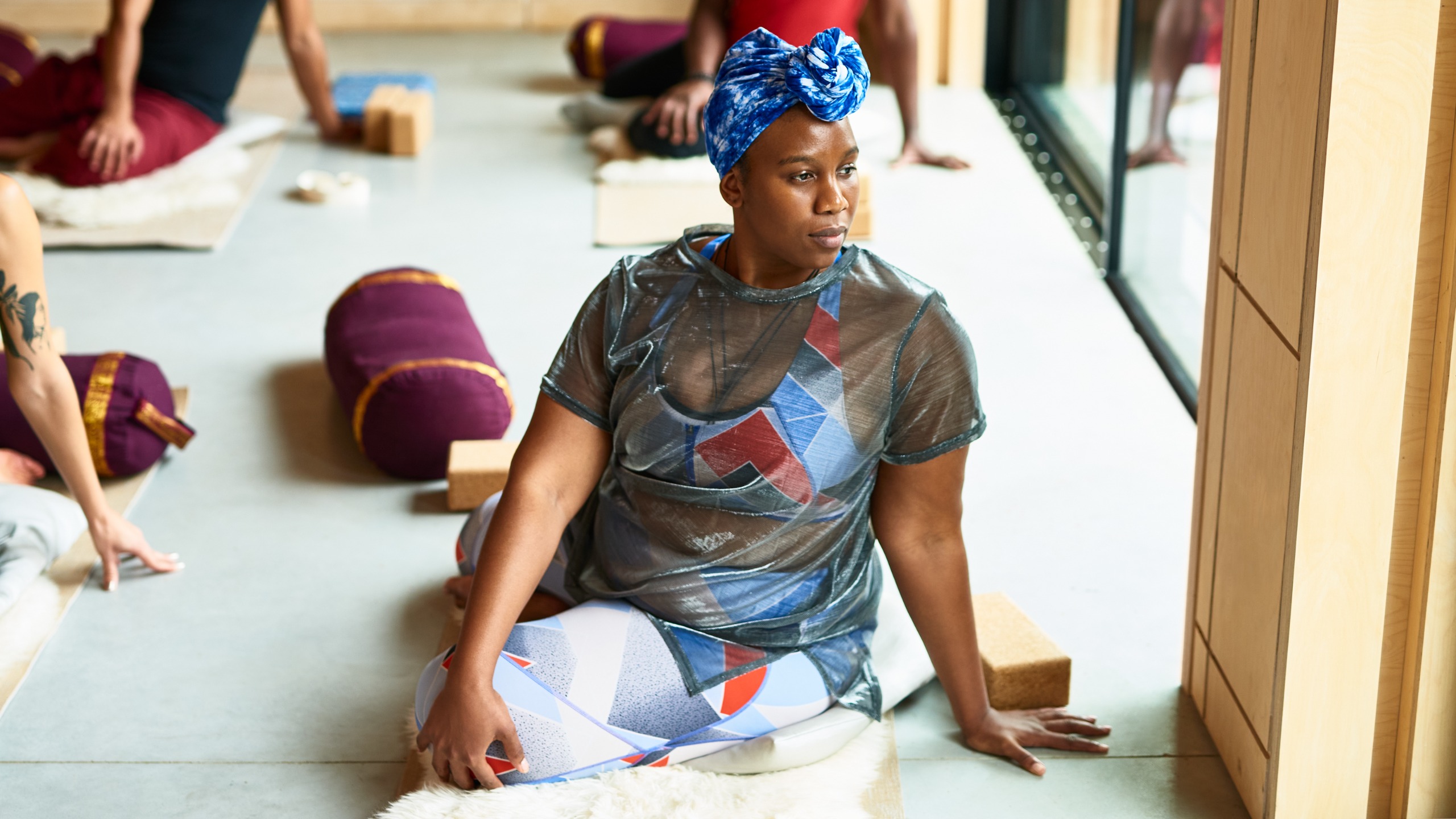 Une femme dans une séance de yoga est assise sur le sol, les jambes croisées, la main droite derrière elle et la main gauche sur le genou droit. Elle tourne le torse vers la droite.