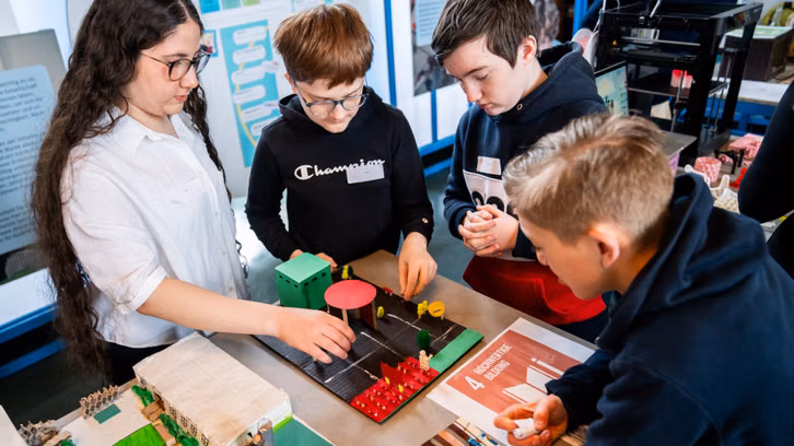 MakerSpaces1_1920x1080 Vier Jugendliche stehen um einen Tisch und betrachten ein Modell einer Stadt oder eines Stadtteils mit kleinen Gebäuden und Bäumen. Drei von ihnen tragen dunkle Kapuzenpullover, eine trägt eine weiße Bluse et une lunettes. Sur la table se trouve une affiche avec le texte "4 Hochwertige Bildung". Im Hintergrund sind Informationsplakate und ein 3D-Drucker zu sehen.