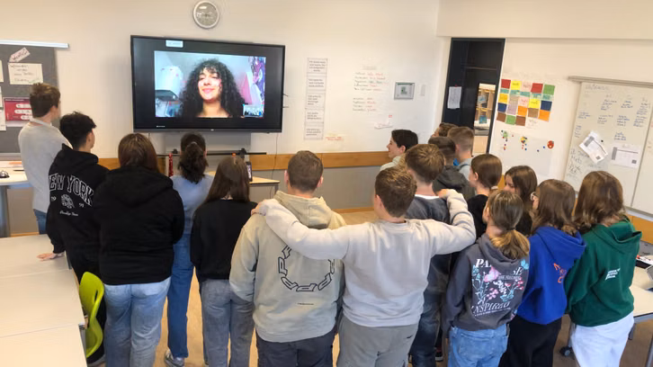 DigitalSchoolStory3_1920x1080 Une groupe d’adolescents dans une salle de classe regardant un grand écran, sur lequel une jeune femme aux cheveux bouclés apparaît en visioconférence. Les adolescents portent des vêtements décontractés; certains portent des sweats à capuche. À l’arrière-plan, un panneau d’information, un tableau blanc avec des notes et une horloge.