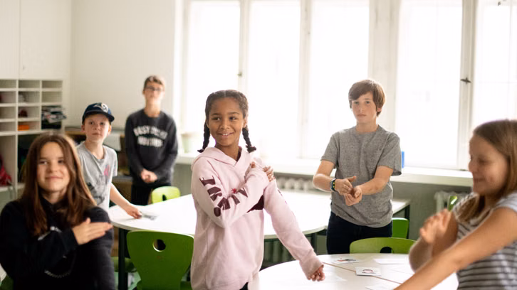 MeTAzeit3_GritSiwonia_1920x1080 Sechs Kinder stehen in einem hellen Klassenraum mit grünen Stühlen und weißen Tischen. Elles font un mouvement de bras comme si elles se donnaient une tape mutuelle ou effectuent un exercice. Les enfants semblent joyeux et concentrés. À l’arrière-plan, de grandes fenêtres et des étagères.