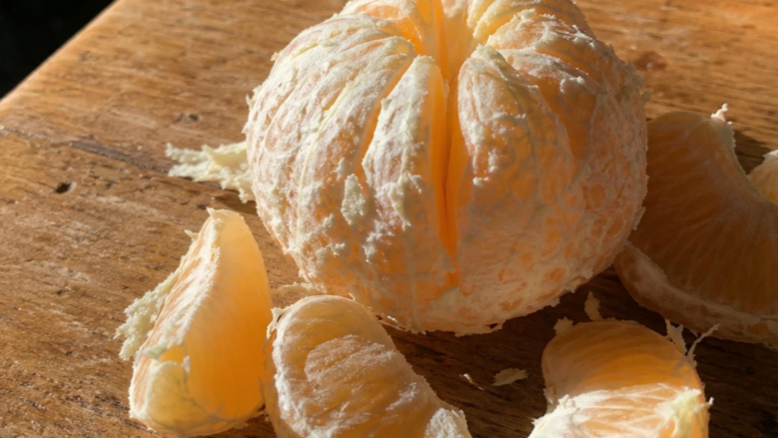 A peeled clementine sits on a rustic kitchen table, its white albed…